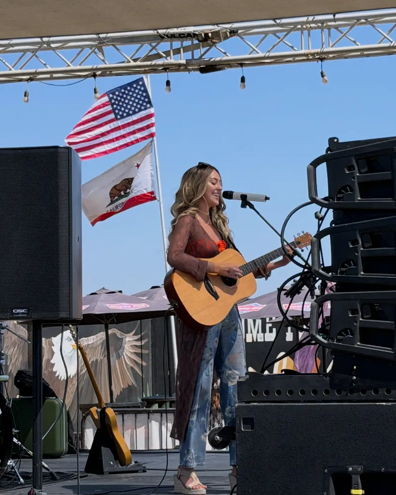 Bella Popa playing acoustic guitar on an outdoor festival stage beneath American and California flags