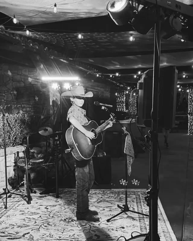 Young performer in a cowboy hat playing guitar on the open mic stage, in black and white